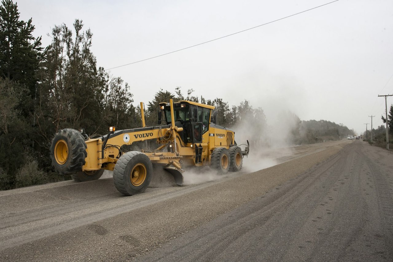 A motor grader levelling a gravel road, throwing dust off the blade as it cuts the running surface