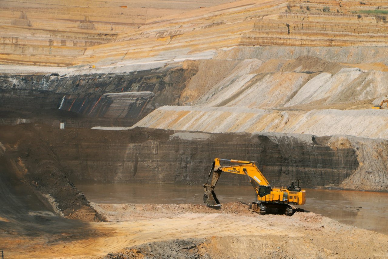 A large open-pit mine worked in benches, with a hydraulic excavator standing on the floor of the cut below stripped overburden